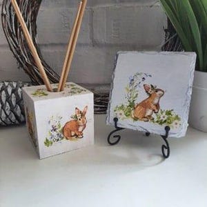 May include: A wooden cube with a painted image of a brown rabbit and flowers, used as a diffuser stand with wooden sticks, and a slate coaster with a painted image of a brown rabbit and flowers.