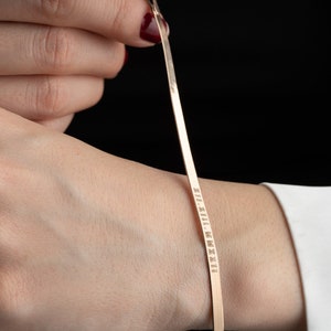 Puede incluir: Una pulsera plana de color dorado con números romanos grabados. La pulsera la sostiene una persona, mostrando su delicado diseño y el texto. La pulsera es una pieza de joyería sencilla y elegante.