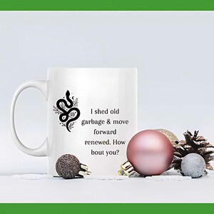 Könnte beinhalten: Eine weiße Keramiktasse mit einem schwarzen Schlangendesign und dem Text "I shed old garbage & move forward renewed. How bout you?"