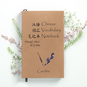May include: A brown leather notebook with the text "Chinese Vocabulary Notebook" and "Hanyu cihui bi ji ben" in Chinese and English. The notebook also has a blue and black ink pen illustration and the name "Caroline" written on the front.