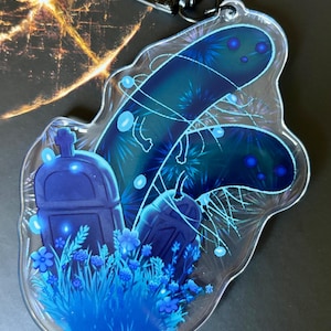 May include: A clear acrylic keychain with a dark blue and teal design. The keychain features a graveyard scene with two tombstones, flowers, and a glowing, abstract design. A silver-colored clasp is attached to the top.