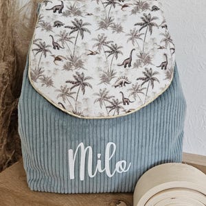 Puede incluir: Mochila de pana azul verdoso con solapa estampada con palmeras y dinosaurios. El nombre "Milo" está escrito en blanco. También se ve un juguete de arcoíris de madera.