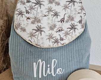 Op de afbeelding: Een lichtblauwe corduroy rugzak met een klep met een patroon van palmbomen en dinosaurussen. De naam "Milo" staat in witte letters. Er is ook een houten regenboog speelgoed te zien.