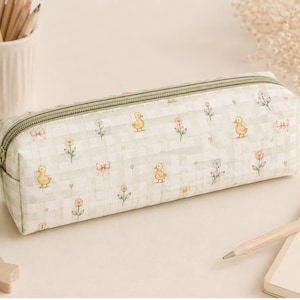 May include: A rectangular pencil case featuring a light green and white gingham pattern. The fabric is decorated with small illustrations of yellow ducklings, pink bows, and various flowers. It includes a matching green zipper closure on top.