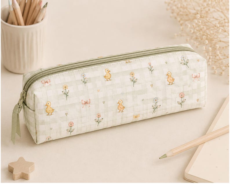 May include: A rectangular pencil case featuring a light green and white gingham pattern. The fabric is decorated with small illustrations of yellow ducklings, pink bows, and various flowers. It includes a matching green zipper closure on top.