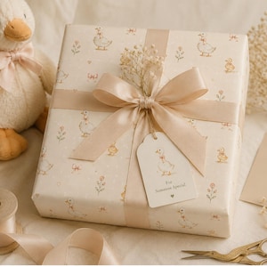 May include: A square gift box wrapped in cream paper with a repeating pattern of small ducks and flowers, tied with a beige satin ribbon. A gift tag attached to the ribbon reads "For Someone Special" with a small heart icon below the text.