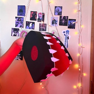 May include: A black, circular, box-like object with a red interior and a row of light pink, triangular teeth-like shapes along the opening. The object features a textured red circular center on its side and is constructed from flat panels.