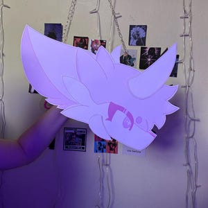 May include: A light purple, flat, stylized character head cutout with pointed ears and a tufted mane. The design features dark purple line art details, including a prominent eye marking and a small snout, presented as a two-dimensional graphic piece.