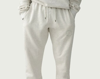 Puede incluir: Pantalones deportivos grises claros con cintura con cordón y puños elásticos. Los pantalones tienen un pequeño logotipo circular en la pierna izquierda. El modelo lleva una sudadera gris claro a juego y zapatillas blancas.