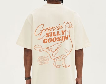 Puede incluir: Camiseta color crema con un gran gráfico naranja de un ganso y las palabras "Groovin' and Silly Goosin'" y "Silly Goose Club". La camiseta tiene mangas cortas y un corte holgado.
