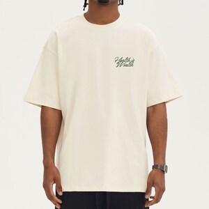 Puede incluir: Camiseta de manga corta color crema con el texto "Health is Wealth" en fuente cursiva. La camiseta tiene un corte holgado y cuello redondo. La tela parece ser un material suave y transpirable.