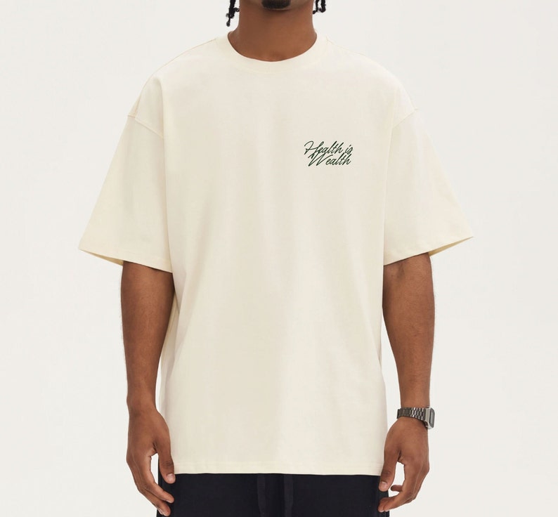 Puede incluir: Camiseta de manga corta color crema con el texto "Health is Wealth" en fuente cursiva. La camiseta tiene un corte holgado y cuello redondo. La tela parece ser un material suave y transpirable.