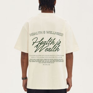 Puede incluir: Camiseta color crema con el texto "Health & Wellness" y "Health is Wealth" en verde oscuro. La camiseta tambi&eacute;n presenta una lista de t&eacute;rminos relacionados con la salud. Corte casual y holgado.