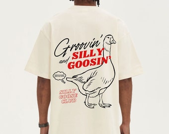 Puede incluir: Camiseta color crema con un gran gráfico de un ganso y las palabras "Groovin' and Silly Goosin'" en negro y rojo. La frase "Silly Goose Club" también está impresa en la camiseta.