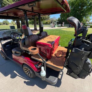 May include: A red golf cart with a canopy, brown leather seats, and a cooler. The cart has a wooden platform and a black golf bag holder. The wheels are a bronze color.