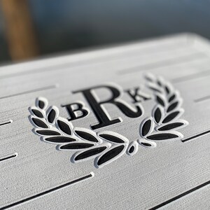 May include: Close-up of a white, textured surface with horizontal lines. The surface features a black laurel wreath design with the letters "B" and "R" and "K" in a bold, sans-serif font. The design is likely for a personalized item.