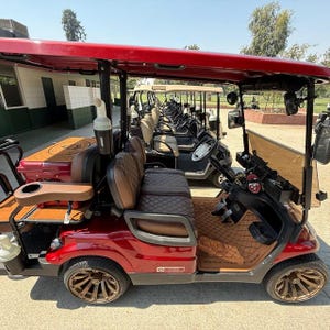 May include: A row of red golf carts with brown seats and a red canopy are parked on a paved surface. The golf carts have brown and gold wheels. The golf carts are parked in a line.