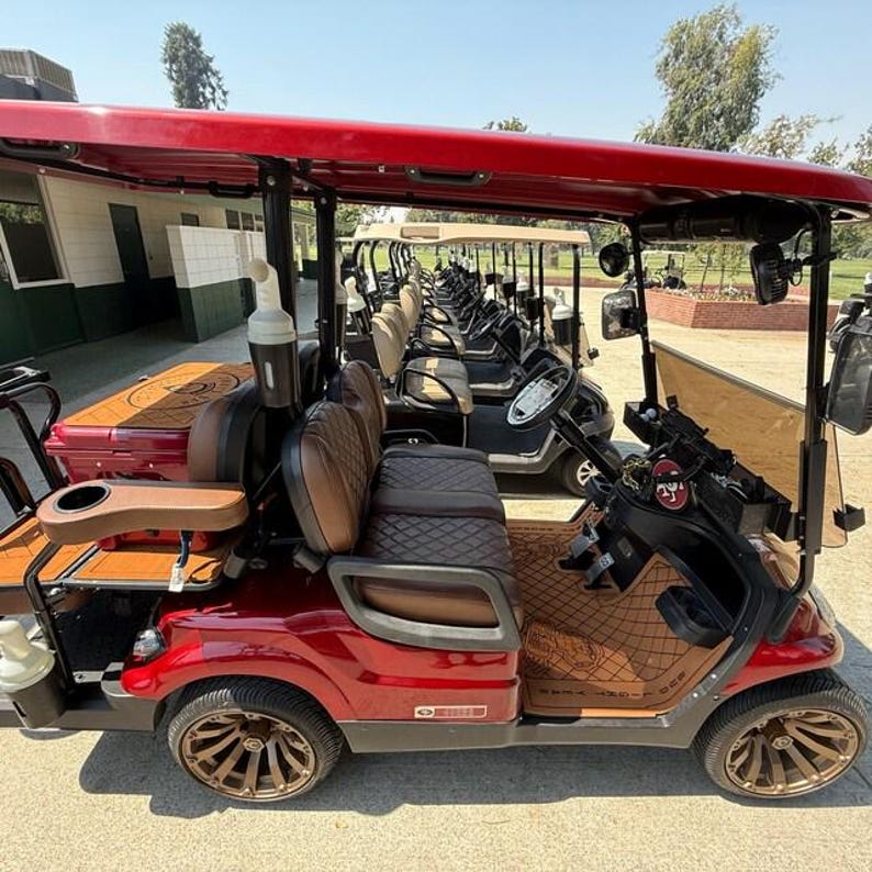 May include: A row of red golf carts with brown seats and a red canopy are parked on a paved surface. The golf carts have brown and gold wheels. The golf carts are parked in a line.