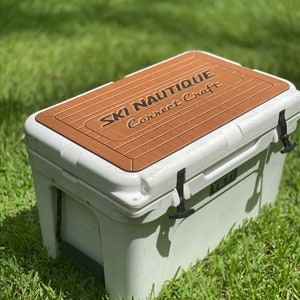 Puede incluir: Nevera blanca Yeti con una tapa de aspecto de madera marr&oacute;n con el texto "SKI NAUTIQUE Correct Craft".