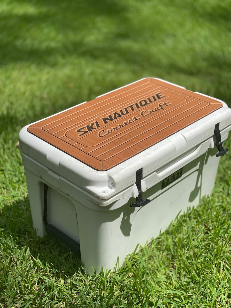 Puede incluir: Nevera blanca Yeti con una tapa de aspecto de madera marr&oacute;n con el texto "SKI NAUTIQUE Correct Craft".