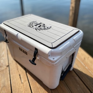 May include: White cooler with a gray top featuring a decorative design. The cooler has black latches and a handle. The cooler is sitting on a wooden dock. The cooler is approximately 24 inches long, 16 inches wide, and 16 inches tall.