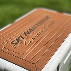 Puede incluir: Una tapa de nevera marr&oacute;n y negra con el texto "SKI NAUTIQUE Correct Craft" impreso en ella.