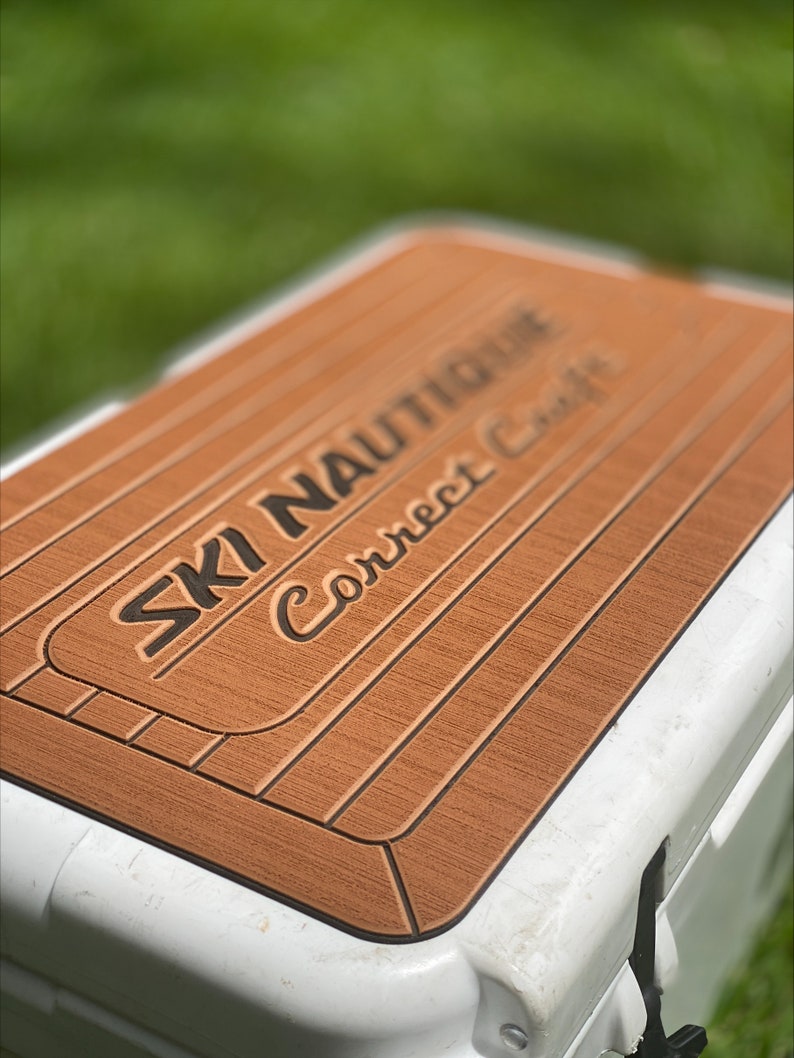 Puede incluir: Una tapa de nevera marr&oacute;n y negra con el texto "SKI NAUTIQUE Correct Craft" impreso en ella.