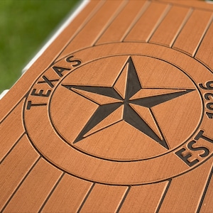 May include: Close-up of a brown wooden surface with a carved Texas star emblem. The star is black and centered within a circle, with the word "TEXAS" above and "EST. 1836" below. The wood has a grooved texture.