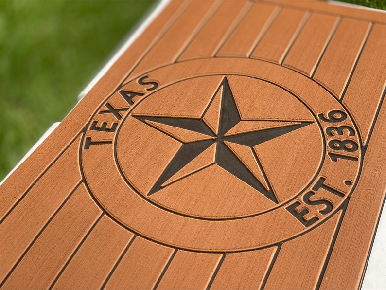 May include: Close-up of a brown wooden surface with a carved Texas star emblem. The star is black and centered within a circle, with the word "TEXAS" above and "EST. 1836" below. The wood has a grooved texture.