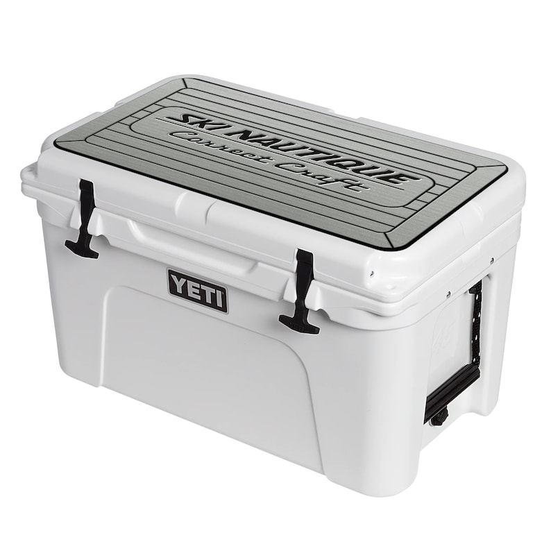 Puede incluir: Nevera blanca Yeti con tapa negra y un logotipo gris de Ski Nautique Correct Craft en la parte superior. La nevera tiene un logotipo negro de Yeti en el lateral.