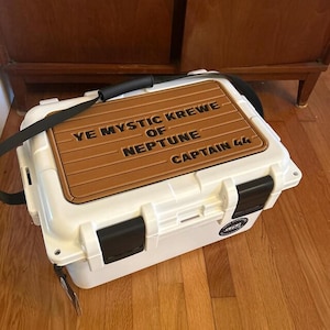 May include: A white cooler with black latches and a brown top with the text "YE MYSTIC KREWE OF NEPTUNE CAPTAIN 44". The cooler has a black shoulder strap and a small sticker on the side.
