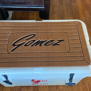 May include: A white cooler with a brown, textured top featuring the name "Gomez" in black script. The cooler has black latches and a red graphic with text. A durable outdoor storage solution.