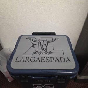 May include: A navy blue and gray cooler with a black handle. The cooler lid features a longhorn skull graphic with the text "XL LARGAESPADA" and "We Are Laughin" in a stylized font. The cooler has the "YETI" logo.