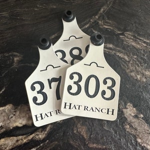 May include: Three white ear tags with black numbers and the text "HAT RANCH". Each tag has a black plastic piece at the top. The tags are stacked on a dark, textured surface.