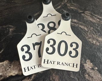Op de afbeelding: Drie witte oormerken met zwarte cijfers en de tekst "HAT RANCH". Elk label heeft een zwart plastic stuk aan de bovenkant. De labels zijn gestapeld op een donker, getextureerd oppervlak.