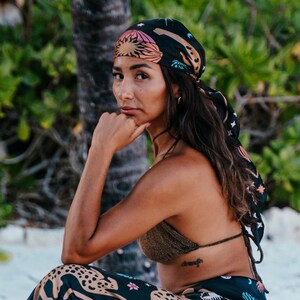 Puede incluir: Una mujer con un pañuelo negro con un diseño celestial colorido y una falda a juego. La falda presenta un estampado de leopardo y un patrón floral. Lleva un top de bikini dorado.