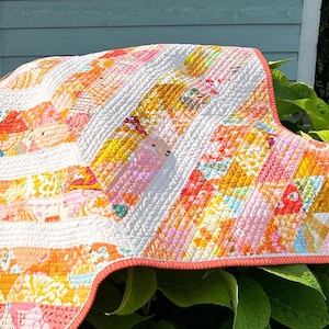 May include: A quilted blanket featuring a patchwork design of various floral and geometric patterns in shades of orange, yellow, pink, and white. The fabric is stitched with a dense, wavy line texture and finished with a solid orange border trim.