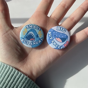 May include: Two blue and white shark themed button badges. The first badge says "Small but Angry" and features a shark with an open mouth. The second badge says "Big but Sweet" and features a shark with a closed mouth and a pink heart.
