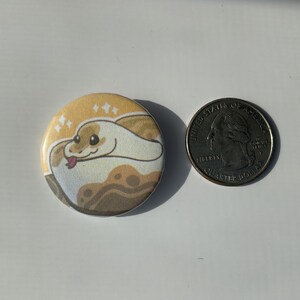 May include: A round button with a cartoon illustration of a snake with a light brown and yellow pattern. The snake is curled up and has a pink tongue sticking out. The background is a light yellow with white sparkles.