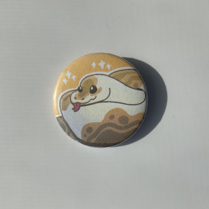 May include: A round button with a cartoon illustration of a white and brown snake with a pink tongue. The snake is curled up on a yellow background with white stars.