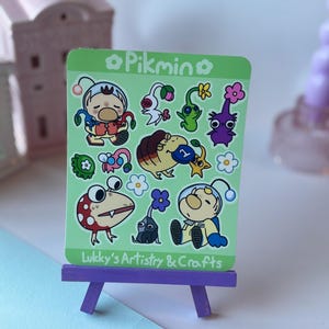 May include: A green sticker sheet featuring various cartoon Pikmin characters. The sticker sheet is displayed on a small purple easel. The text "Pikmin" and "Lukky's Artistry & Crafts" are visible on the sticker sheet.