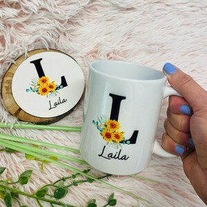 Könnte beinhalten: Weiße Keramiktasse und passender Untersetzer mit dem schwarzen Buchstaben "L" mit Sonnenblumen- und Grünakzenten. Der Name "Laila" ist darunter aufgedruckt. Der Untersetzer ist aus Holz.