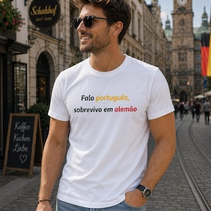 Könnte beinhalten: Weißes T-Shirt mit dem Text "Falo português, sobrevivo em alemão" in Schwarz, Gelb und Rot. Das T-Shirt wird von einer Person getragen, die eine Sonnenbrille und eine blaue Jeans trägt. Die Person steht auf einer Kopfsteinpflasterstraße.
