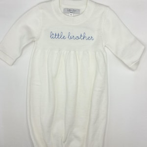 May include: A white baby gown with long sleeves. The words "little brother" are embroidered in blue script across the chest. A small tag is visible near the neckline. The gown is made of a soft, knit fabric.