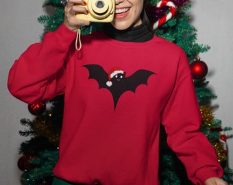 Könnte beinhalten: Ein roter Weihnachtspullover mit einem schwarzen Fledermaus-Motiv, das einen Weihnachtsmann-Hut trägt. Der Pullover wird von einer Person getragen, die vor einem Weihnachtsbaum steht.
