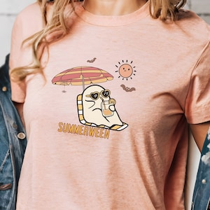 Puede incluir: Una camiseta rosa con un fantasma de dibujos animados que lleva gafas de sol y sostiene una bebida. El fantasma está sentado en una silla de playa bajo una sombrilla. El texto "SUMMERWEEN" está impreso debajo del fantasma.