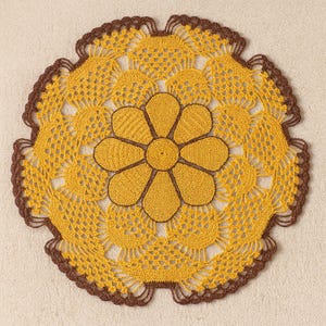 Könnte beinhalten: Handgefertigte, gelbe Häkelspitze mit braunem, gewelltem Rand und einem großen Blumendesign in der Mitte. Die Blume ist ebenfalls gelb mit braunen Details. Die Spitze ist kreisförmig und hat ein offenes, durchbrochenes Muster.