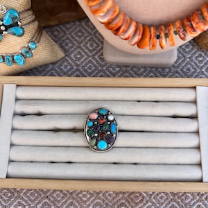 May include: An oval silver ring featuring a cluster of small, multi-colored cabochon gemstones including turquoise, coral, and other earth-toned stones set in a decorative twisted wire frame, displayed on a beige velvet ring tray insert.