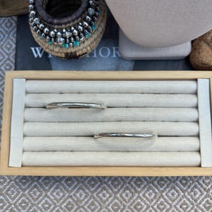 May include: Two silver bangles with engraved geometric patterns resting in a rectangular wooden display tray with cream-colored velvet slots. The top bangle features a repeating dash design, while the bottom bangle displays a series of small circles.