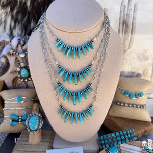 May include: A multi-strand silver necklace featuring four cascading tiers of turquoise heishi beads and small silver spacer beads. The necklace is displayed on a beige bust, surrounded by various turquoise rings, bracelets, and earrings.